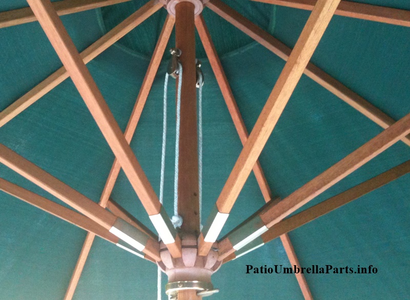 two brass market umbrella pulleys attached to a central wood brown pole hold ropes which help raise and lower the green umbrella canopy shade